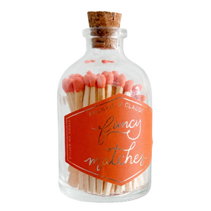 A clear glass bottle with a cork stopper filled with wooden matchsticks and a bright orange label that reads 'Fancy matches' by 'Frankie & Claude'.