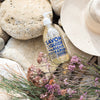Glass Liquid Soap bottle labeled "Savon Liquide Marseille Extra pur, Mediterranee" on top of rocks next to a sun hat and wildflowers.